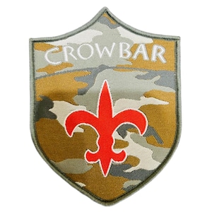 May include: Shield-shaped patch with a camouflage pattern in shades of green, brown, and gray. The word "CROWBAR" is embroidered in white across the top. A red fleur-de-lis emblem is centered below the text. The patch has a green border.