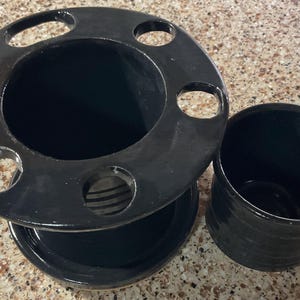 May include: Black ceramic toothbrush holder with a circular top featuring six holes, a central compartment, and a matching small cup. The set is glossy and appears to be handmade.