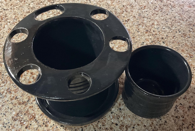 May include: Black ceramic toothbrush holder with a circular top featuring six holes, a central compartment, and a matching small cup. The set is glossy and appears to be handmade.