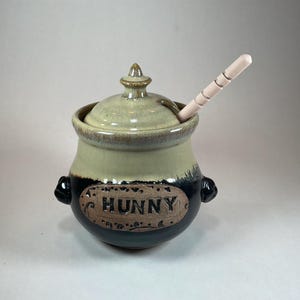 May include: A ceramic honey pot with a two-tone glaze, featuring a dark black base and a light beige top. The front has an oval plaque with the word HUNNY embossed in black. It includes a matching lid with a finial and a light wooden honey dipper.