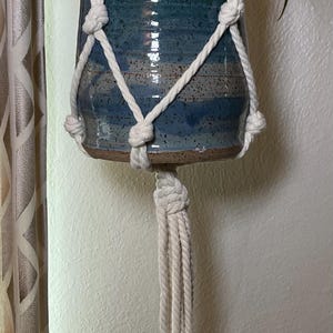 May include: A ceramic plant pot with a blue and green glaze, suspended by a cream-colored macrame plant hanger. The pot holds a green plant with long, slender leaves. The macrame has a knotted design.