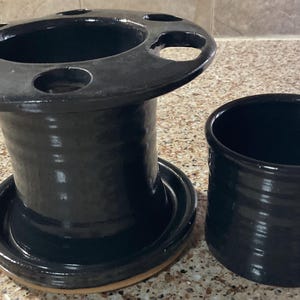 May include: A black ceramic toothbrush holder and a matching tumbler. The toothbrush holder has a wide, circular top with several holes and a cylindrical base. The tumbler is a simple, cylindrical shape. Both items have a glossy finish.