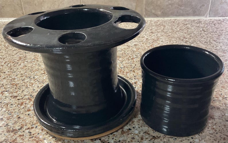 May include: A black ceramic toothbrush holder and a matching tumbler. The toothbrush holder has a wide, circular top with several holes and a cylindrical base. The tumbler is a simple, cylindrical shape. Both items have a glossy finish.