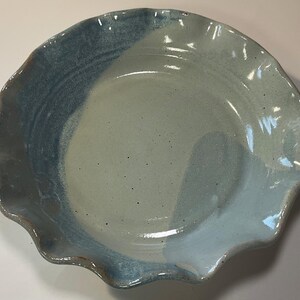 May include: A handmade ceramic bowl with a wavy, ruffled edge. The bowl is glazed in shades of blue and light green, creating a two-tone effect. The interior is a light green, while the rim is a darker blue. The bowl is approximately 10 inches in diameter.