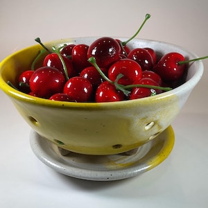 May include: A ceramic colander bowl with a yellow and white speckled glaze, featuring drainage holes along the sides and a matching saucer base. The bowl is filled with numerous glossy, bright red artificial cherries with green stems.