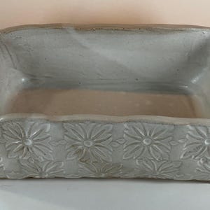 May include: A rectangular ceramic baking dish with a glossy white glaze. The exterior features a repeating embossed floral pattern across the sides and handles. The dish has two integrated handles on the ends and a smooth, open interior basin.