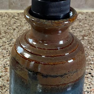 May include: A handcrafted ceramic soap dispenser with a dark brown and blue glaze. The dispenser has a black pump and a textured surface, perfect for a bathroom or kitchen.