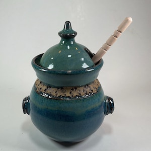 May include: A teal ceramic honey pot with a matching lid and two side handles. The pot features a textured band around the upper body and includes a light-colored wooden honey dipper resting inside the lid opening.