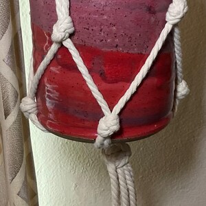 May include: A handmade macrame plant hanger with a red and burgundy ceramic pot. The pot is suspended by a woven, off-white rope. Green plant leaves are visible inside the pot. The hanger is hanging in front of a window.