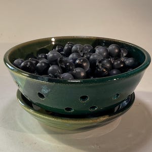 May include: A dark green ceramic berry bowl with a matching saucer base. The bowl features a series of small, circular drainage holes around its lower perimeter and is filled with a collection of small, round, dark purple-black berries.
