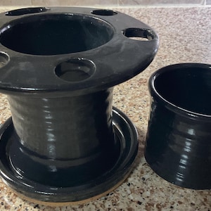 May include: A black ceramic toothbrush holder with multiple holes and a matching cylindrical cup. The holder has a wide, circular top and a round base. The set is glazed with a glossy finish.