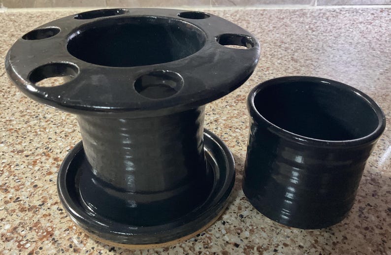 May include: A black ceramic toothbrush holder with multiple holes and a matching cylindrical cup. The holder has a wide, circular top and a round base. The set is glazed with a glossy finish.
