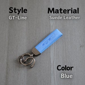 May include: A light blue suede leather keyring with a silver key ring and the text "GT-Line" embossed on the leather. The keyring is on a grey wooden surface.