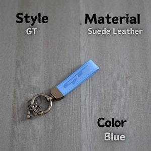 May include: Blue suede leather keyring with a silver metal ring and a GT logo. The keyring is on a wooden surface.