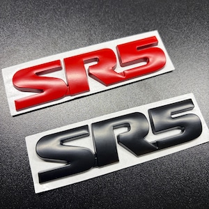 Puede incluir: Dos emblemas SRS, uno rojo y otro negro, sobre un soporte blanco. El emblema rojo es brillante, mientras que el emblema negro tiene un acabado mate. Estos emblemas son probablemente para la personalización automotriz.