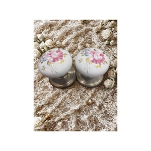 May include: Two white ceramic door knobs with a floral design in pink, blue, and yellow. The knobs have a silver-colored base and are set against a lace-like fabric background. The floral design features roses.