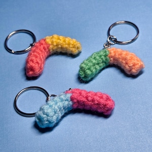 May include: Three colorful, handmade crochet worm keychains. Each worm is a different combination of colors: pink and yellow, green and orange, and blue and pink. The keychains have silver metal rings and are set against a blue background.