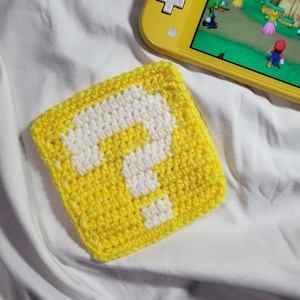 May include: A yellow crocheted square with a white question mark in the center. The square is lying on a white surface. A yellow Nintendo Switch Lite is visible in the background.