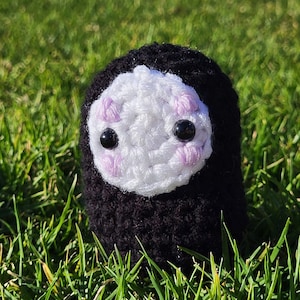 May include: A crocheted plush toy with a black body and a white face. The toy has two black eyes and pink cheeks.