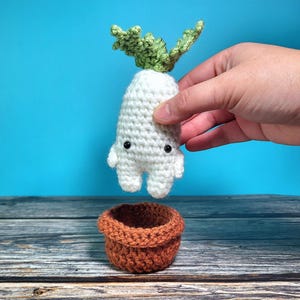 May include: A white crocheted radish with green leaves, with a cute face, is being held above a brown crocheted pot.