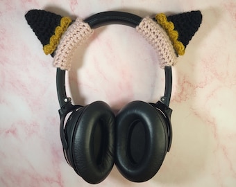 Pode incluir: Auscultadores pretos com capas de orelhas de gato de crochet rosa e preto. As capas das orelhas têm uma guarnição dourada.