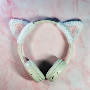 May include: White wireless headphones with pink and white crocheted cat ears. The headphones are on a pink marble background.