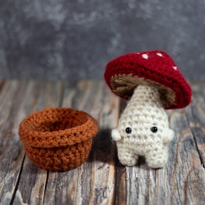 May include: A crocheted mushroom toy with a red and white cap and a white body. The mushroom is standing on a wooden surface next to a brown crocheted pot.