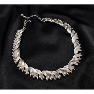 May include: A silver necklace with a leaf-shaped design. The necklace is composed of curved, textured metal elements, creating a layered appearance. The piece is displayed on a black backdrop, accentuating its intricate details.