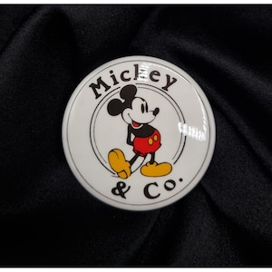 May include: A white circular button featuring Mickey Mouse in a classic pose, wearing red shorts, yellow shoes, and black gloves. The button has the text "Mickey & Co." in a black font, encircled by a black line.
