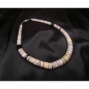 May include: A necklace made of flat, round, natural-colored beads. The beads are primarily white, tan, and brown, with black accents. The necklace has a silver clasp and is displayed on a black background. A unique piece of jewelry.