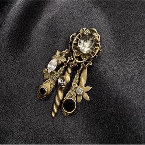 May include: An antique gold-tone brooch with dangling charms. The brooch features a large, oval, clear gemstone at the top, surrounded by an ornate frame. Below, there are several charms with clear and black gemstones, and floral and spiral designs.