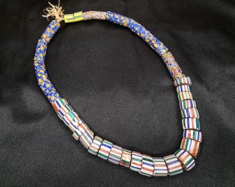 May include: A colorful necklace featuring a variety of beads. The necklace has a blue patterned section, and a section of striped beads in red, green, white, and blue. The necklace is displayed on a black surface.