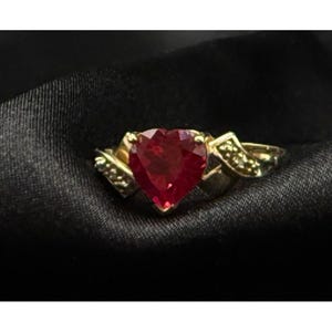 May include: A gold ring featuring a prominent heart-shaped red gemstone at the center. The band is crafted from gold and includes small, clear accent stones embedded along the shoulders on both sides of the central heart-shaped red stone.