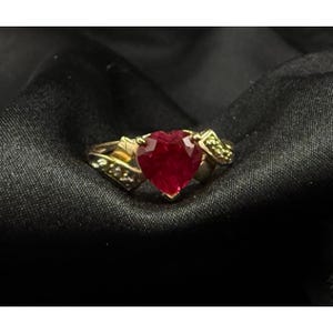 May include: A gold ring featuring a prominent heart-shaped red gemstone at the center. The band is crafted from yellow gold and includes small, clear accent stones embedded along the sides of the setting, framing the central red heart.
