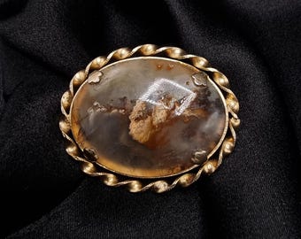 May include: An oval-shaped antique brooch with a gold-toned twisted rope border. The center stone is a translucent, light brown and gray agate with a unique pattern. The brooch is set against a black fabric background.