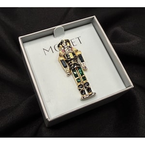 May include: A gold-tone nutcracker brooch featuring black and green enamel accents and clear rhinestones. The brooch is displayed on a light gray card inside a box with the text MONET printed in black letters on the upper portion of the card.