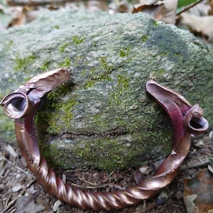 May include: A twisted copper bracelet with a unique design. The bracelet has two decorative ends that resemble animal heads. The bracelet is resting on a mossy rock.