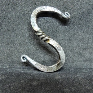 May include: A hand-forged metal letter S with a twisted design. The letter is made of silver-colored metal and has a smooth, polished finish.