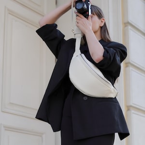 May include: A black blazer and skirt ensemble paired with a white fanny pack. A black camera is held up to take a photo. The outfit is worn in front of a white door.