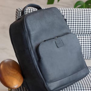 Peut inclure: Un sac à dos en cuir noir avec une poignée supérieure et plusieurs compartiments zippés. Le sac à dos porte le mot "BOPOR" en relief sur la poche avant. Le sac est posé sur une chaise à motifs noir et blanc.