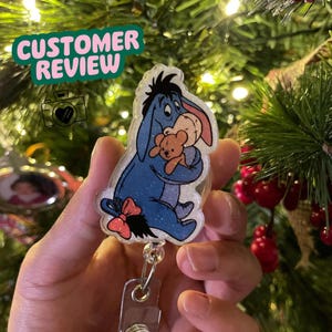 K&ouml;nnte beinhalten: Ein blau glitzernder Ausweis-Rollhalter mit Eeyore aus Winnie the Pooh, der einen Teddyb&auml;ren h&auml;lt. Der Ausweis-Rollhalter hat eine rote Schleife und einen transparenten Ausweishalter. Der Text "CUSTOMER REVIEW" befindet sich in einer gr&uuml;nen Blase.