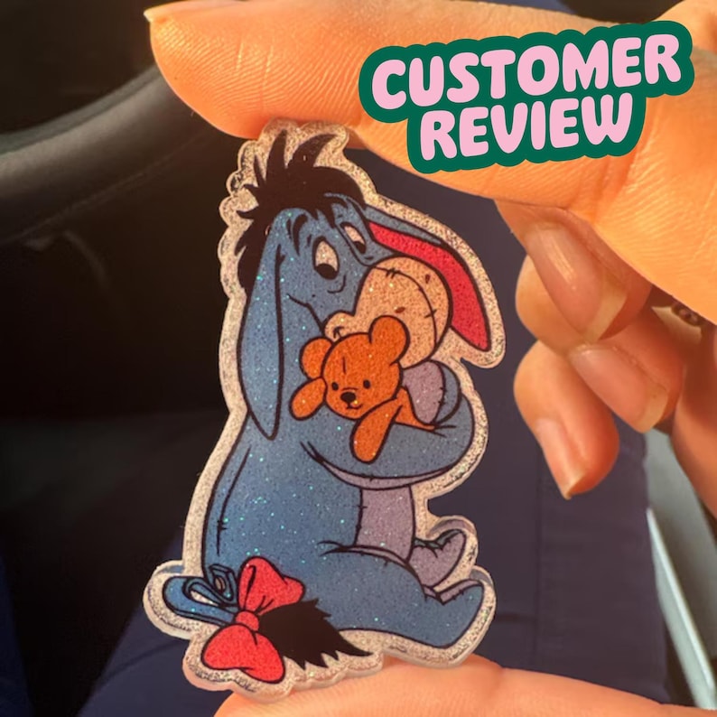 K&ouml;nnte beinhalten: Ein glitzernder Acryl-Pin mit Eeyore aus Winnie Puuh, der einen Teddyb&auml;ren umarmt. Eeyore ist blau mit einer roten Schleife, und der Teddyb&auml;r ist orange. Der Pin hat einen klaren Rand und die Worte "CUSTOMER REVIEW" in einem gr&uuml;nen Banner.