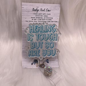 以下が含まれることがあります： 「HEALING IS TOUGH BUT SO ARE YOU」の文字がライトブルーで書かれたバッジリール。リールは透明なプラスチックストラップと、銀色のキラキラした装飾が付いています。リールの後ろのカードにはお手入れ方法が記載されています。