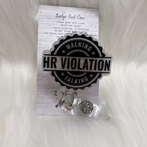 May include: A silver badge reel with a black and white glitter sticker that says "Walking HR Violation Talking". The reel is on a white background.