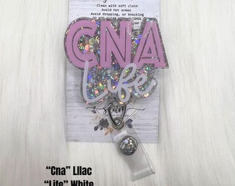 May include: A badge reel featuring a clear resin topper filled with silver galaxy glitter. The topper displays the text CNA in large lilac letters and the word life in white script. A metal clip and clear plastic strap are attached to the base.
