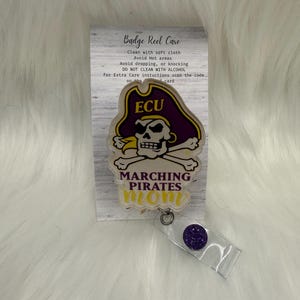 以下が含まれることがあります： 紫と金の海賊帽をかぶったドクロとECUの文字、その下にMARCHING PIRATES momと書かれたバッジリール。バッジリールは、Badge Reel Careと書かれたお手入れ方法が記載された白い台紙に取り付けられています。
