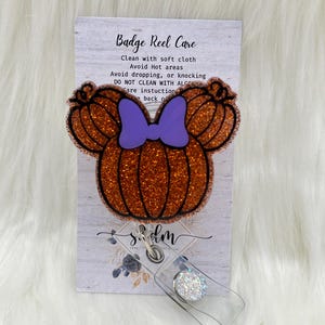 May include: Orange glitter pumpkin badge reel with a purple bow. The reel has a clear plastic strap and a sparkly white button. The card attached to the reel has instructions for caring for the badge reel. The text on the card reads "Badge Reel Care", "Clean with soft cloth", "Avoid Hot areas", "Avoid dropping, or knocking", "DO NOT CLEAN WITH ALCOHOL", "Care instructions", "back of".