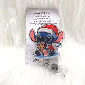 Pode incluir: Um porta-crachá com um personagem de desenho animado azul usando um gorro de Papai Noel, segurando um biscoito e uma xícara de chocolate quente. O porta-crachá tem um cordão retrátil de plástico transparente e um botão decorativo brilhante.