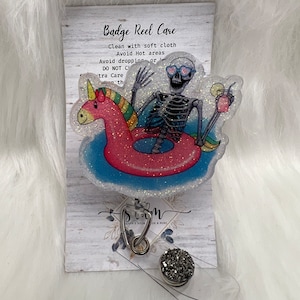 Puede incluir: Un carrete de insignia transparente con un esqueleto cubierto de purpurina montando un flotador de unicornio. El carrete tiene un dije plateado con una piedra brillante. La tarjeta detrás del carrete tiene instrucciones de cuidado: "Badge Reel Care Clean with soft cloth Avoid Hot areas Avoid dropping or DO NOT C tra Care th".