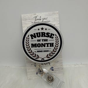 以下が含まれることがあります： 白い背景に黒い文字でNurse of the MonthとJune 2026と書かれた円形のバッジリール。黒い円形の縁取りと2つの月桂樹の葉のデザインが施されています。バッジリールには、金属製のスナップが付いた透明なプラスチック製のストラップが含まれています。